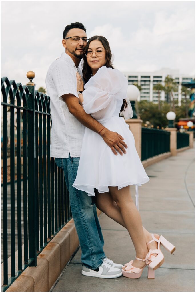 The couple is standing together, smiling as they pose for a photo at Disneyland’s California Adventure. The woman wears a white puffy-sleeved dress, and the man is dressed casually in a white shirt and jeans.