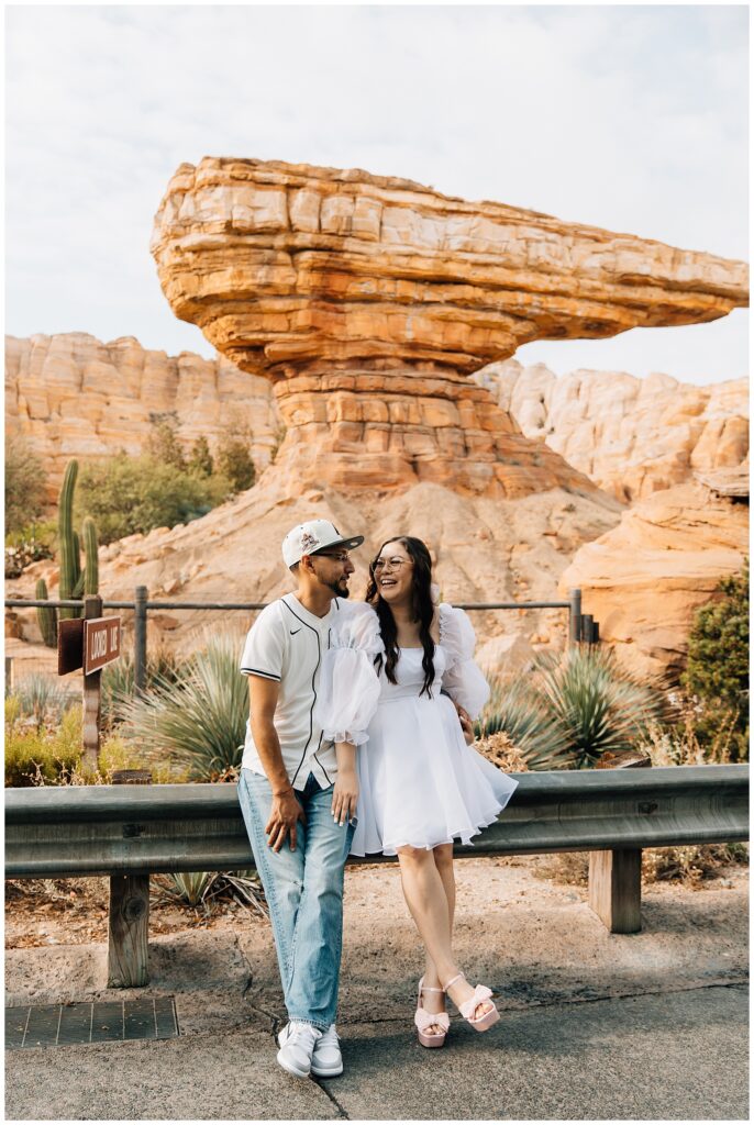 The couple sits near the iconic Radiator Springs rock formation at Disneyland’s California Adventure, sharing a light-hearted moment. The woman playfully kicks her legs while the man looks lovingly at her.