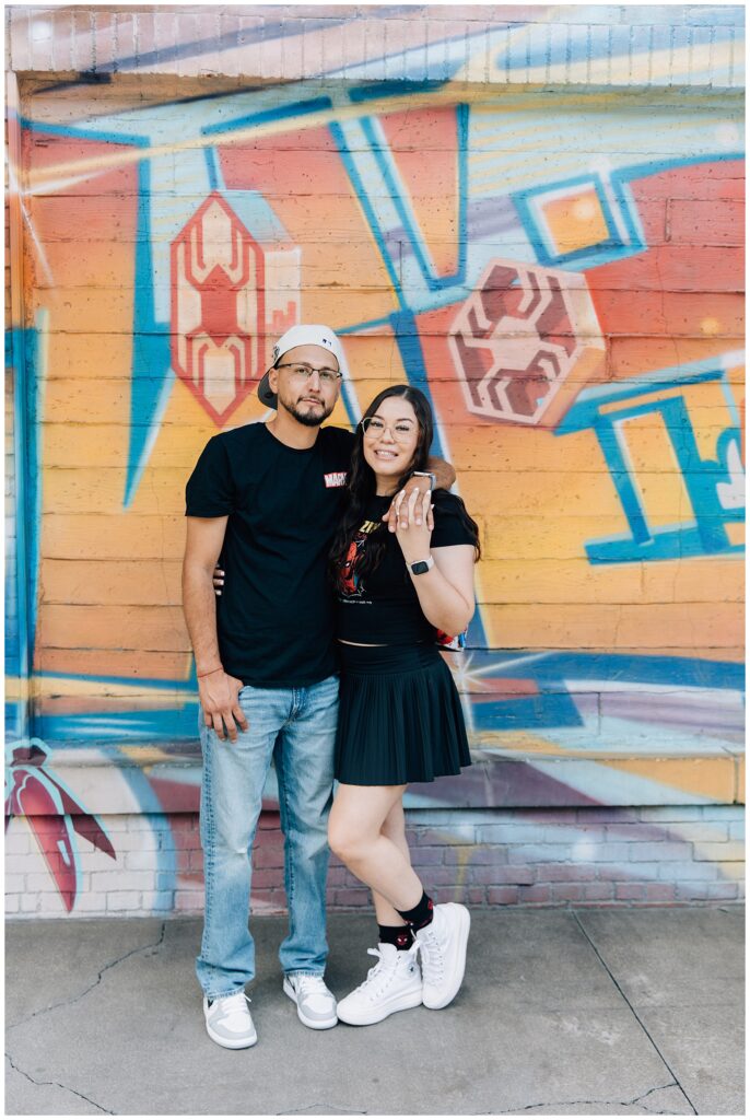 The couple stands in front of a brightly colored mural in Disneyland's Avengers Campus, the woman proudly displaying her engagement ring. They are dressed in casual Marvel-themed clothing, enjoying the fun atmosphere.