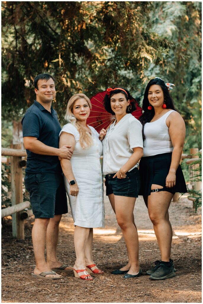 Two women and one man stand close together in a wooded area, with one of the women holding a red parasol. The group is smiling, standing on a forest path, surrounded by trees.