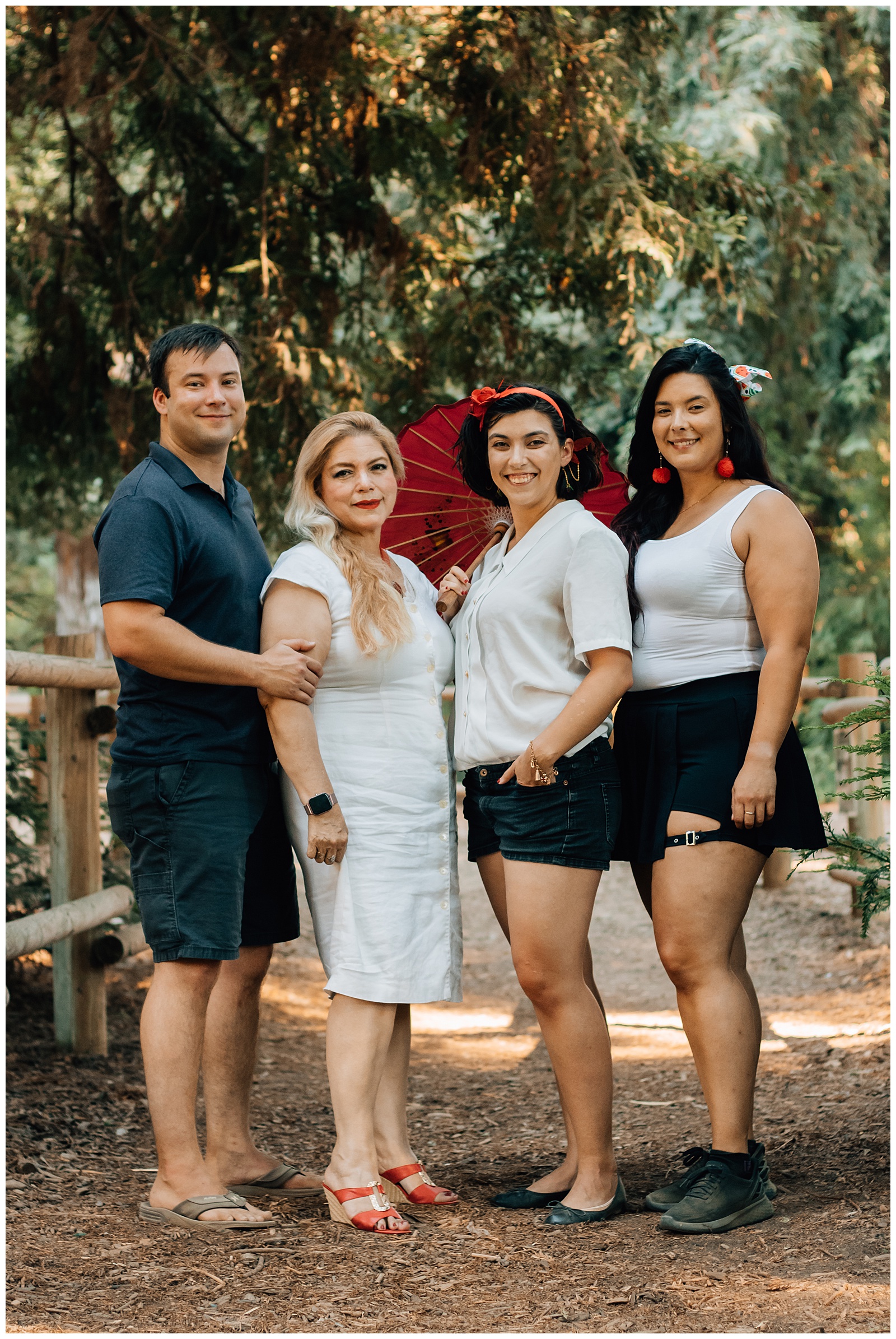 Two women and one man stand close together in a wooded area, with one of the women holding a red parasol. The group is smiling, standing on a forest path, surrounded by trees.