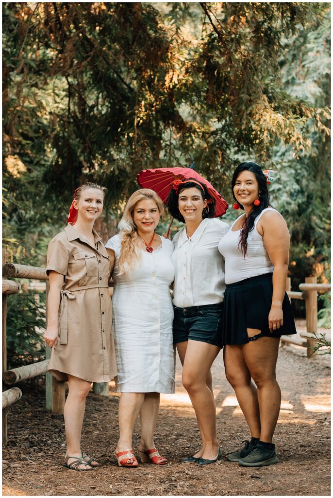 A group of four women stands together on a wooded path, with one of the women holding a red parasol. They are all smiling at the camera, surrounded by trees and soft, warm lighting.