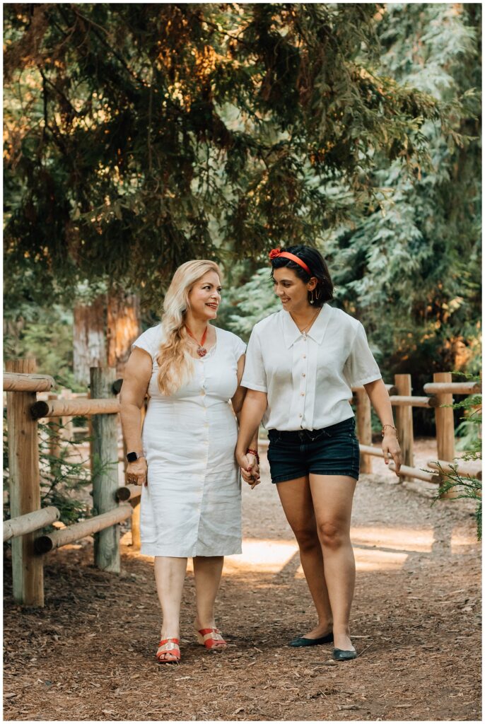 Two women, one wearing shorts and the other in a white dress, are walking hand in hand along a forest path. They are smiling at each other, with trees and sunlight surrounding them.