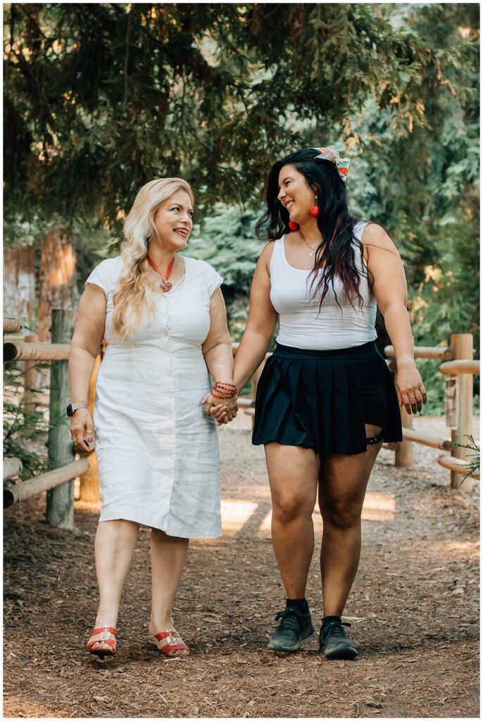 Two women walk hand in hand, looking at each other and smiling as they stroll down a path in the woods. The atmosphere is light and natural, with greenery all around them.