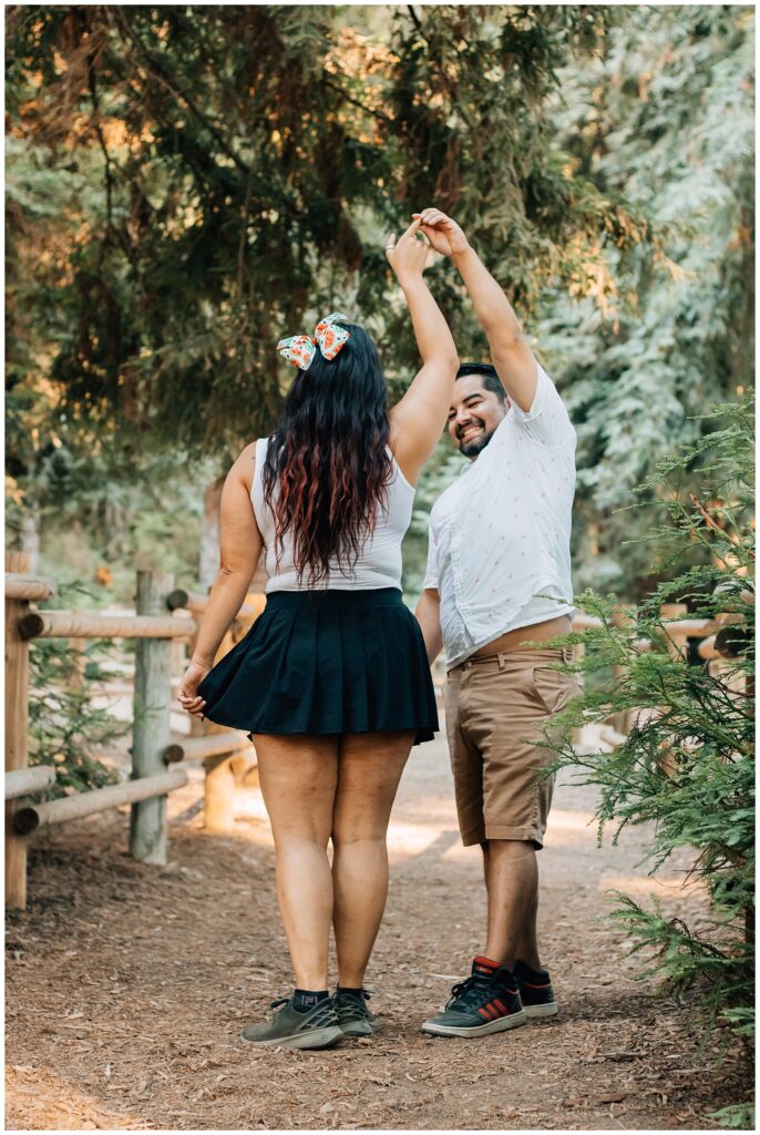 A man and woman are enjoying a lighthearted moment as he twirls her in a playful dance. They are standing on a forest path, surrounded by trees and soft natural light.