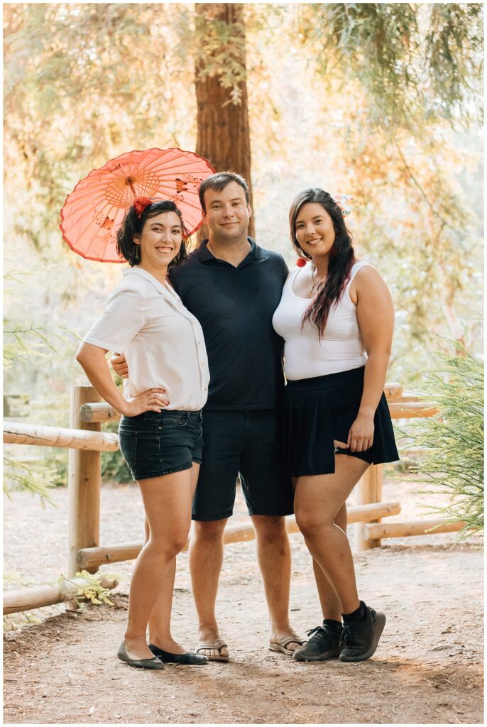 Two women and one man stand together in a wooded area, with one woman holding a red parasol. They are dressed casually, smiling at the camera, with tall trees and soft lighting in the background.