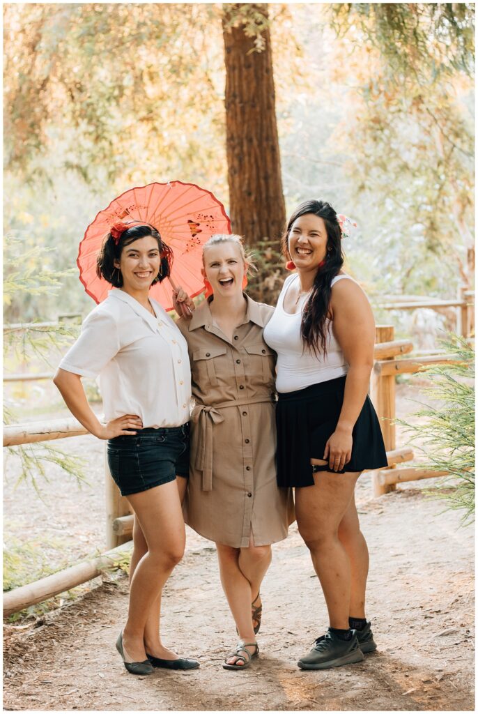 Three women stand together, smiling and laughing, with one woman holding a bright red parasol behind them. They are in a wooded area, with dappled sunlight shining through the trees.