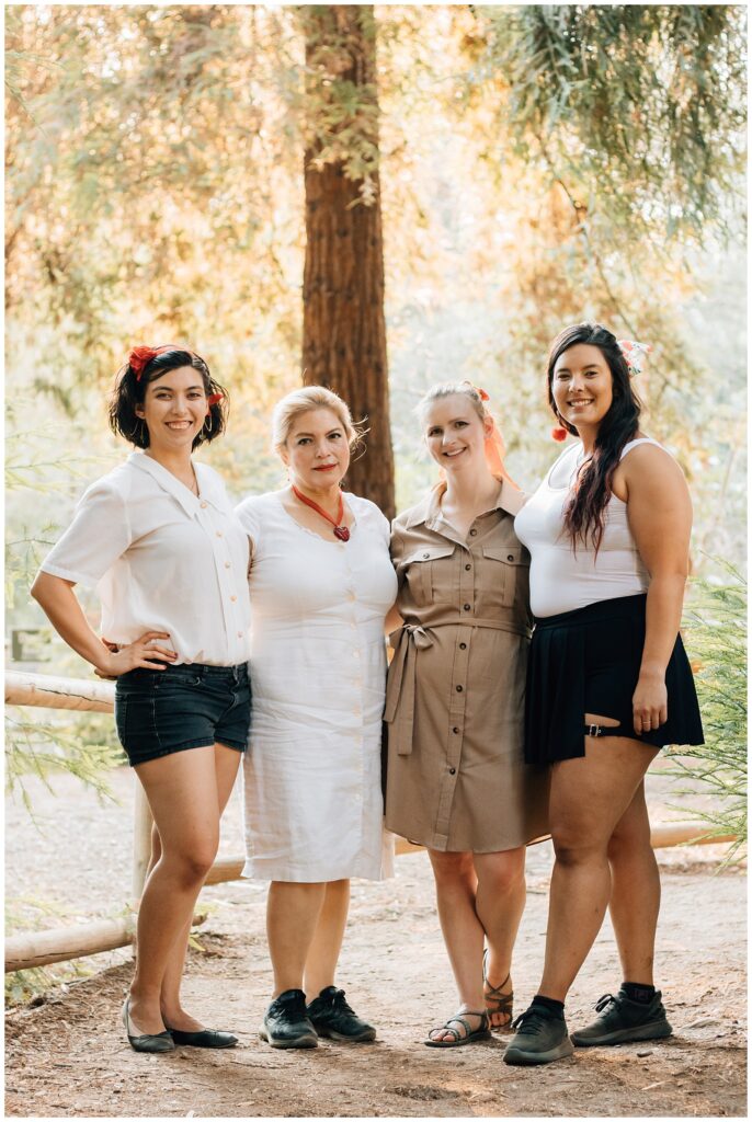 A group of four women standing side by side in a wooded area. They are smiling at the camera, dressed casually in light-colored outfits, with trees and soft sunlight filtering through the background.