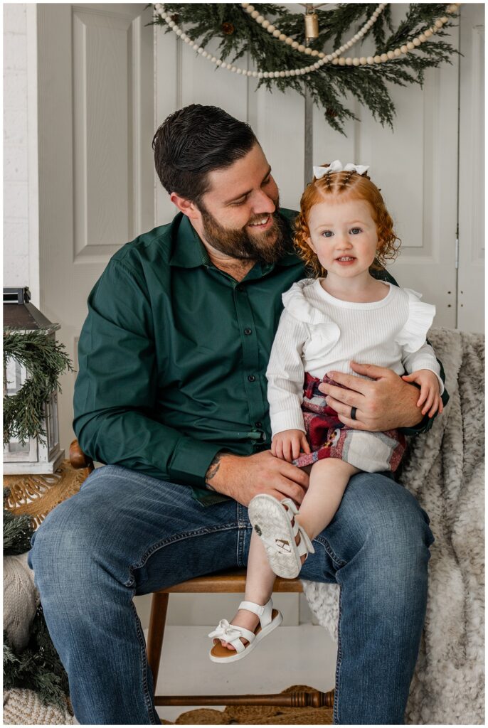 A father with a dark beard, wearing a forest green shirt, sits on a wooden chair holding his young red-haired daughter on his lap. The girl, smiling slightly, wears a white long-sleeved ruffled top, a plaid red skirt, and white sandals with bows. Festive greenery and holiday decorations hang in the background.