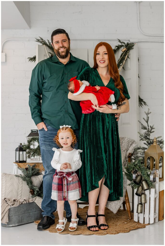 A family of four stands together in front of a holiday-decorated backdrop. The father, wearing a green shirt and jeans, stands to the left; the mother, with long red hair and wearing a green velvet dress, cradles a newborn baby in a red Santa outfit. The red-haired toddler stands in front of her parents, wearing a plaid skirt and white sandals.