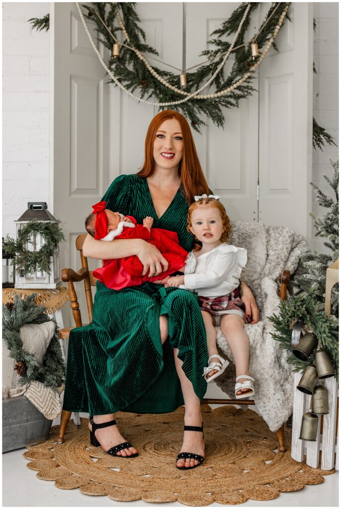 A mother with long red hair sits in a wooden chair wearing a green velvet dress, holding a sleeping newborn dressed in a red Santa outfit. Beside her sits her red-haired toddler daughter, dressed in a white ruffled top and red plaid skirt, looking directly at the camera. Festive greenery and blankets surround the scene.