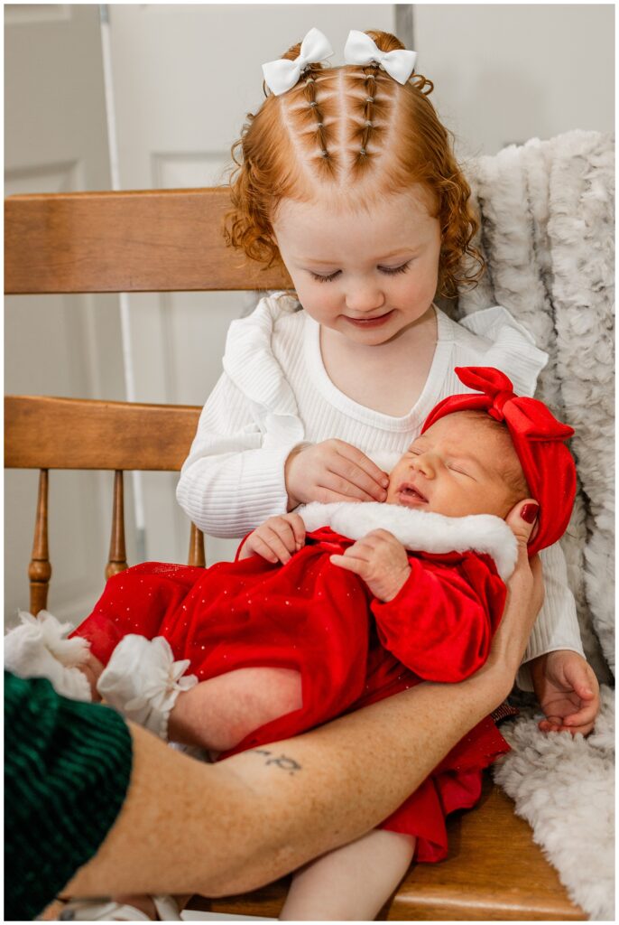 A close-up of a toddler girl with red hair styled into small braids, adorned with white bows, sitting on a wooden chair. She gazes lovingly at a sleeping newborn baby in her arms, who is dressed in a red velvet Santa outfit with a matching red bow.