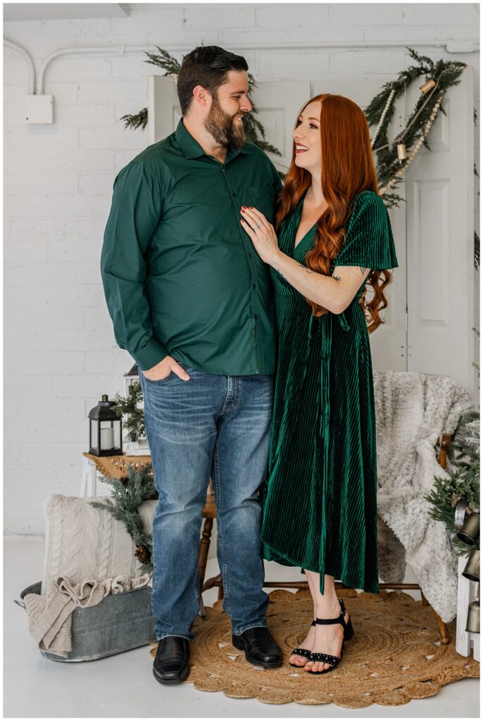 A couple stands side-by-side, smiling warmly at each other. The man wears a dark green shirt and jeans, while the woman, with long red hair, wears a green velvet dress. Festive holiday decorations frame the backdrop, with greenery, string lights, and cozy blankets completing the scene.