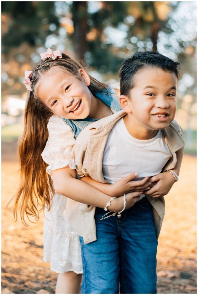 A playful moment between a young boy and girl as the girl hugs her brother from behind, both laughing joyfully. They are outdoors with blurred trees and warm lighting in the background.