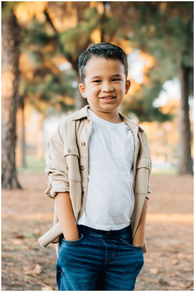A young boy poses confidently with his hands in his pockets, wearing a beige shirt over a white T-shirt and jeans. He stands in a park with trees and golden light in the background.