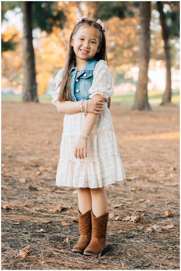 A young girl in a white ruffled dress and denim vest stands with her arms crossed, smiling brightly. She wears brown cowboy boots and pink hair bows, with autumn leaves scattered at her feet.