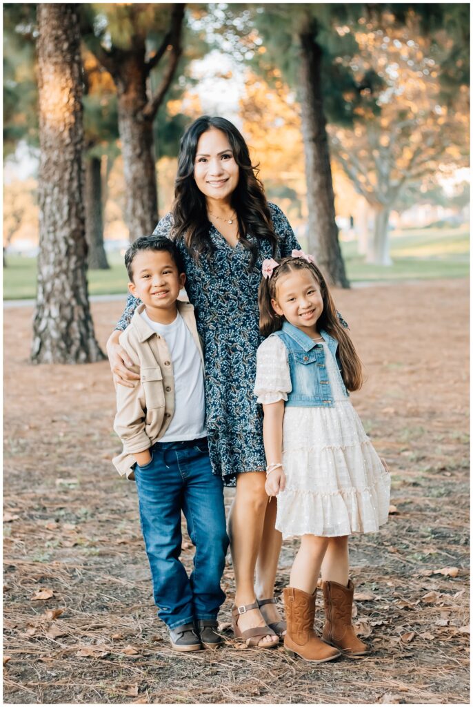 A mother poses with her two children, who stand on either side of her, all smiling warmly. The setting is a park with trees and a golden-hour glow in the background.