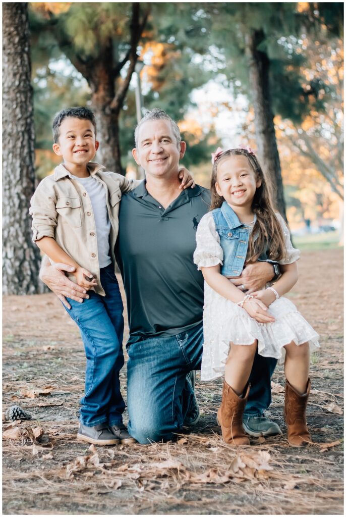A father kneels in a park, holding his two children close on either side. The boy rests an arm on his father’s shoulder, and the girl sits on her father’s knee, both smiling happily.