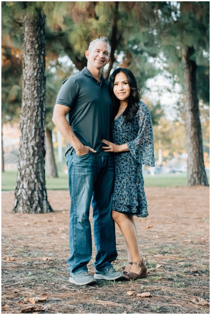 A couple stands together in a serene park setting. The man wears a gray polo and jeans, while the woman wears a flowing blue-patterned dress. They stand close, smiling softly at the camera.