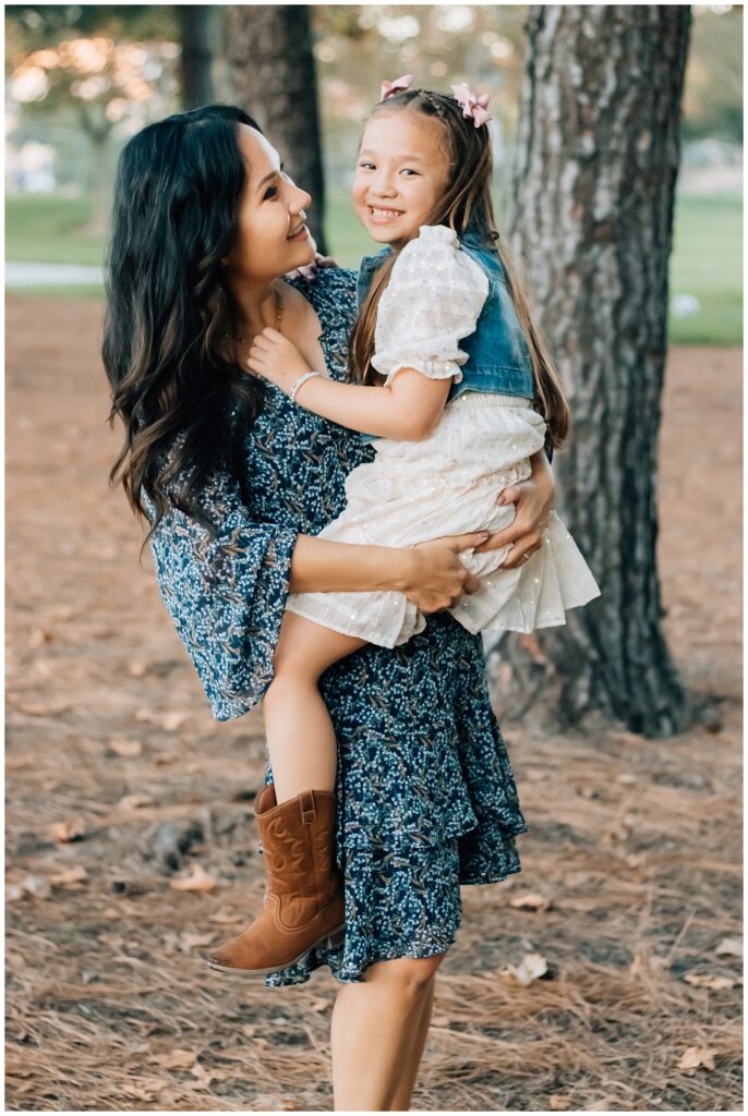 A mother lifts her young daughter in her arms while they share a loving gaze. The girl wears a white dress and cowboy boots, and the park background is bathed in warm, natural light.