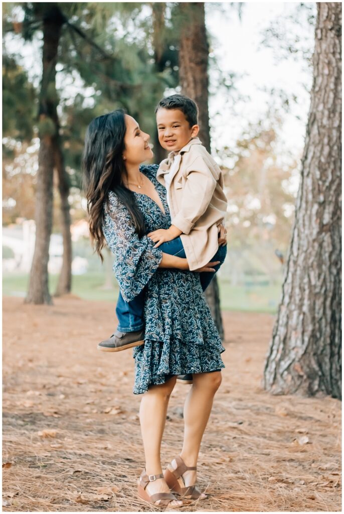 A mother holds her young son close, both smiling at each other warmly. The boy wears a beige shirt and jeans, while the mother wears a blue-patterned dress, surrounded by a park's tall trees.