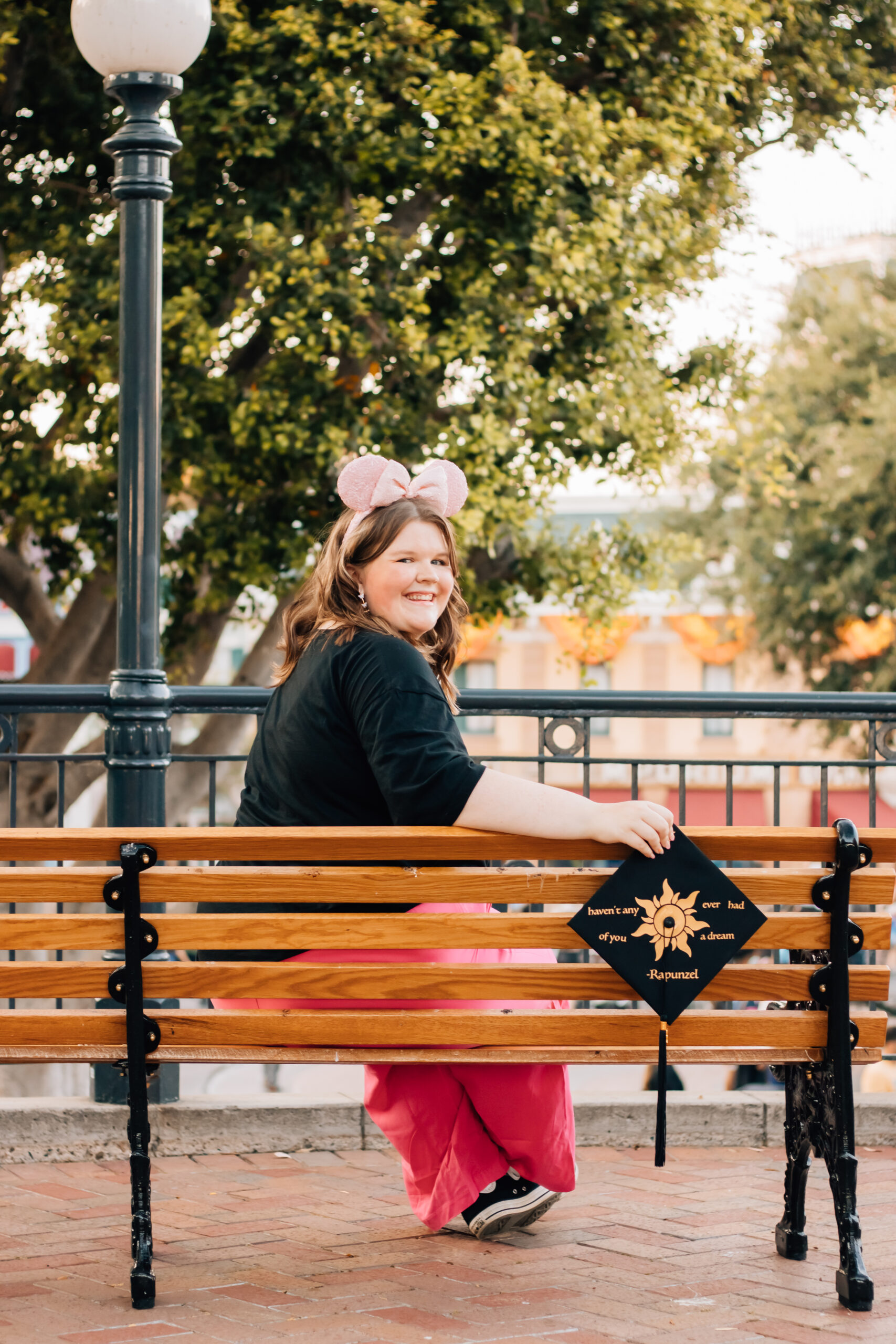 Disneyland graduate wearing pink Minnie ears and pink pants smiling on bench while holding decorated Rapunzel-inspired graduation cap