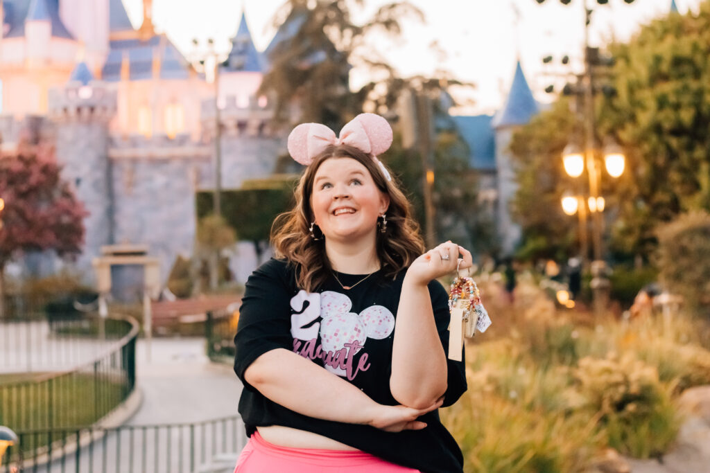 Disneyland graduate holding class keychain in front of Sleeping Beauty Castle at sunset, wearing 2025 graduate shirt and pink Minnie ears