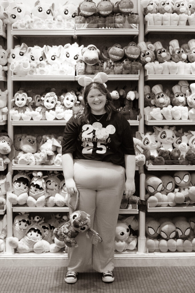 Disneyland graduate in 2025 shirt posing with Stitch plush toy in front of colorful wall of Disney stuffed animals