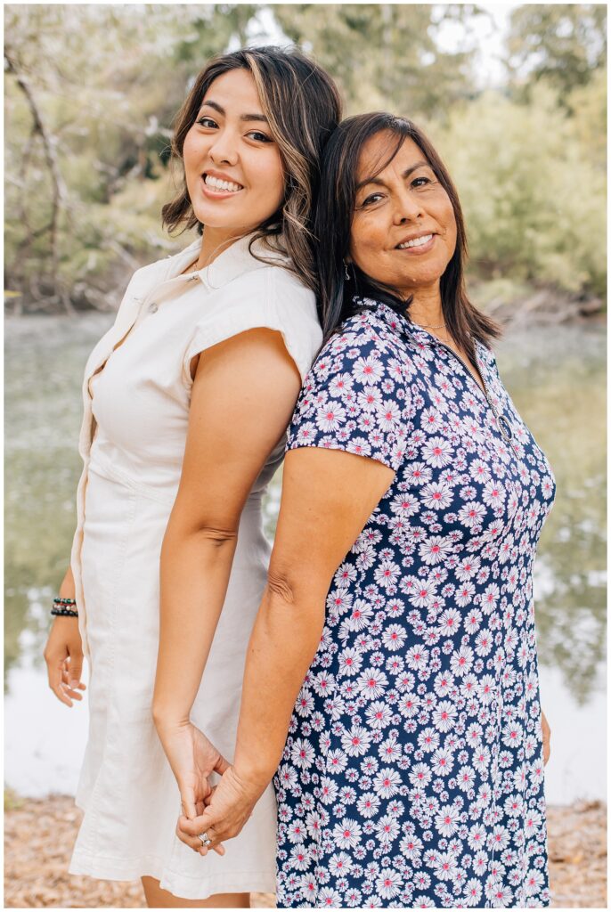 Mother and daughter standing back-to-back near a peaceful pond, holding hands and smiling.