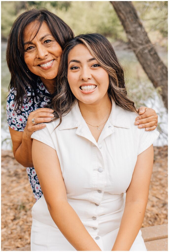 Mother standing behind her seated daughter with her hands on her shoulders, both smiling warmly at the camera.
