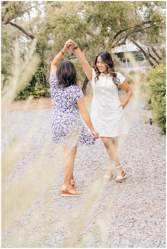 Mother and daughter dancing together on a gravel path surrounded by trees, with the daughter twirling under her mom's raised hand.