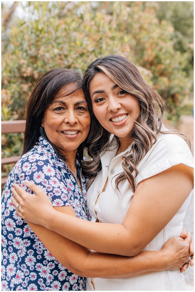 Mother and daughter hugging and smiling at the camera during an outdoor photo session, surrounded by greenery.