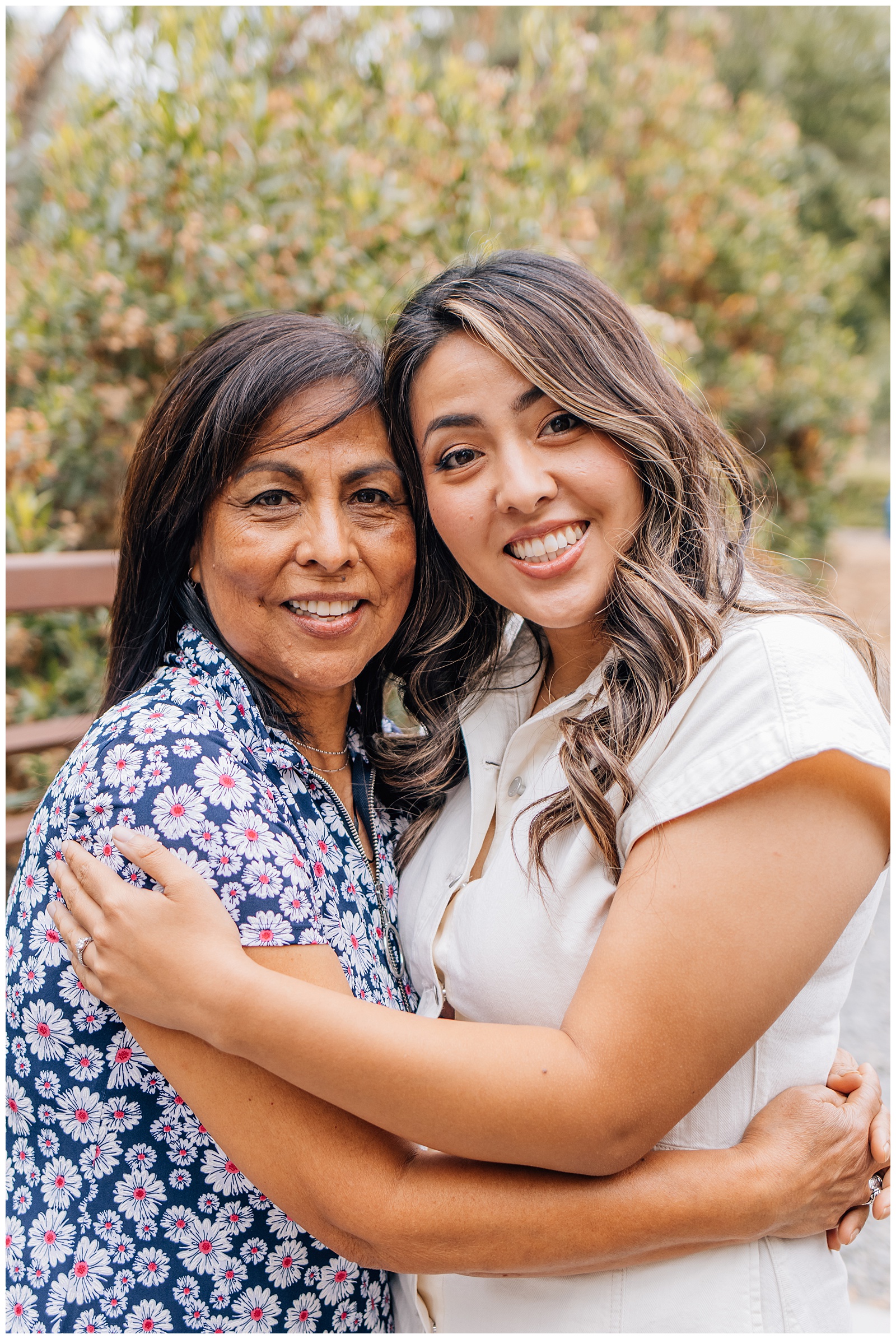 Mother and daughter hugging and smiling at the camera during an outdoor photo session, surrounded by greenery.