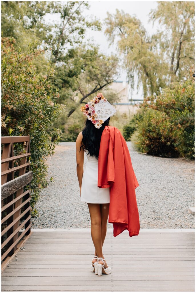 A graduate stands on a wooden bridge, facing away from the camera, wearing a strapless white dress and holding a red graduation gown draped over one shoulder. Her graduation cap is decorated with flowers and reads, “Every storm ends in a bloom.”