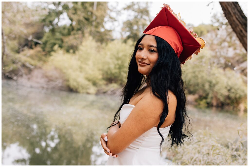A smiling graduate in a red cap and strapless white dress poses by a calm river with trees in the background. Her arms are crossed, and her long black hair flows over her shoulders.
