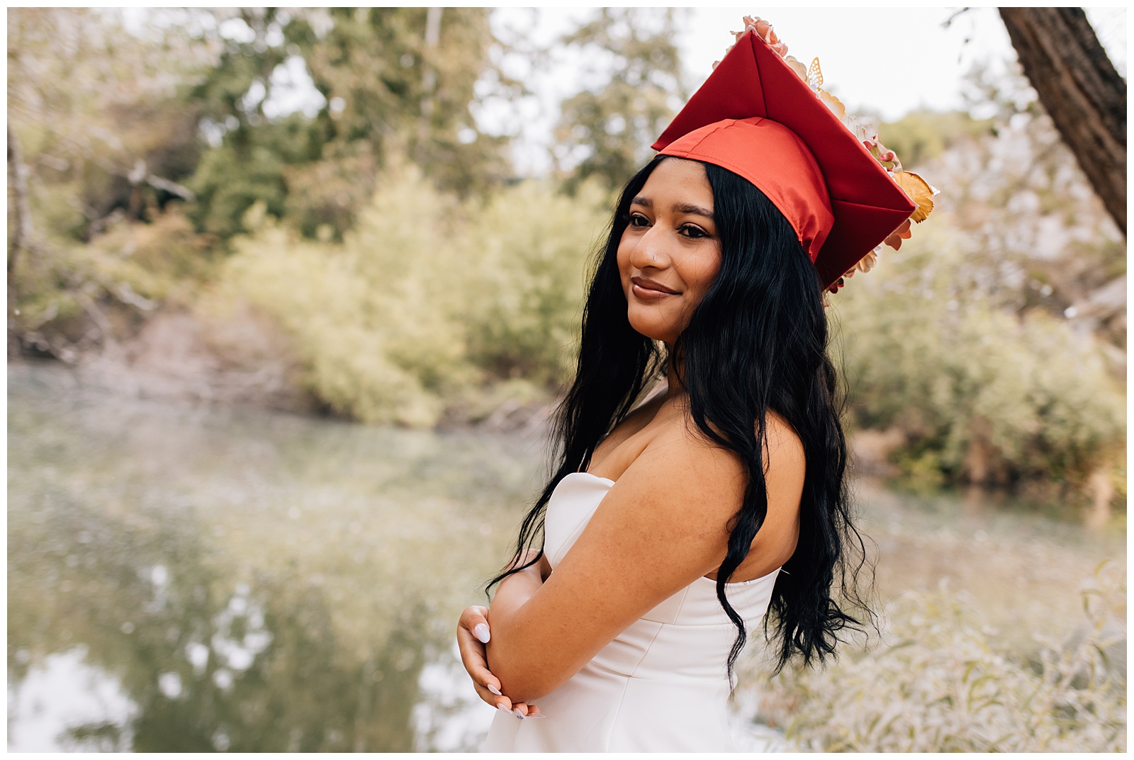 A smiling graduate in a red cap and strapless white dress poses by a calm river with trees in the background. Her arms are crossed, and her long black hair flows over her shoulders.