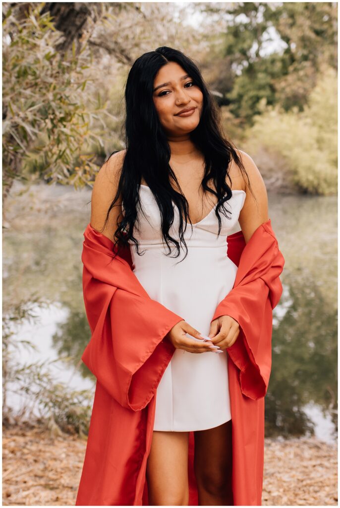A graduate wearing a white dress and red gown draped over her arms stands in front of a tree-lined river, smiling gently at the camera.