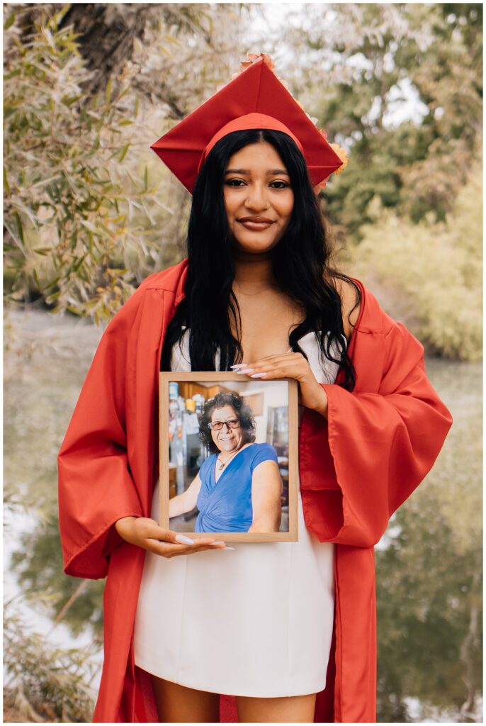 A graduate in a red cap and gown smiles while holding a framed photo of a smiling woman in a blue shirt, honoring a loved one. The background features a peaceful wooded area and river.